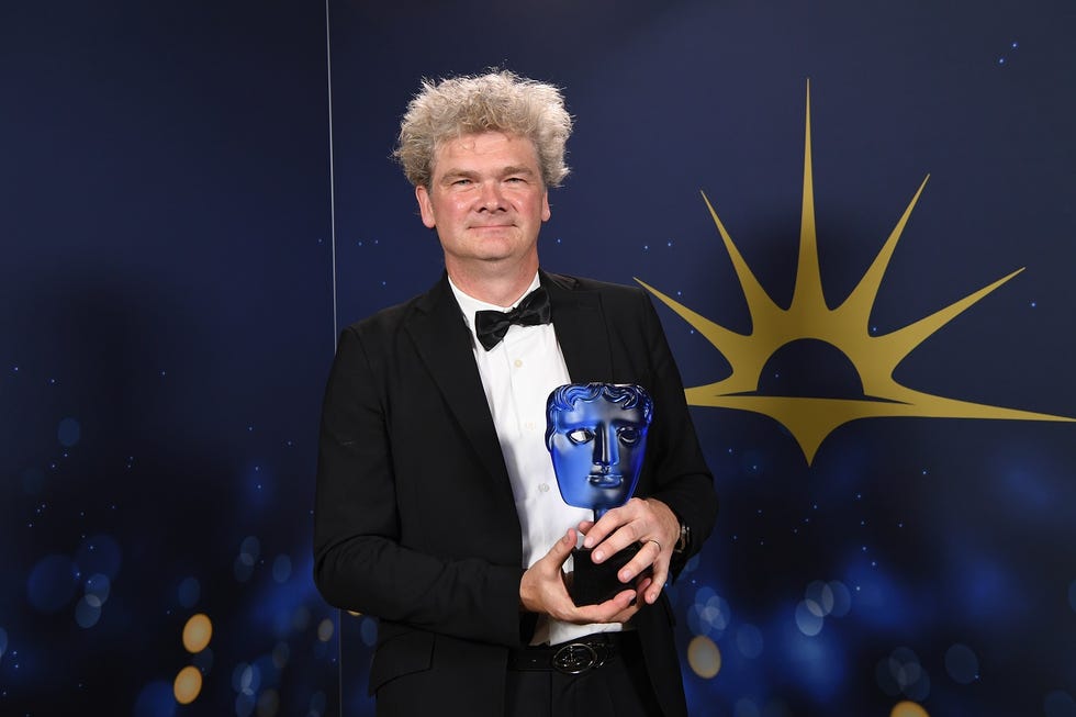 simon farnaby at the 2023 bafta television awards, simon is looking at the camera while wearing a tuxedo and holding a blue bafta award