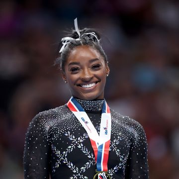 simone biles smiles at the camera, she wears a black leotard with gemstones, a white hair ribbon, and a gold medal with a red, white, and blue lanyard