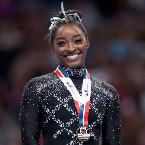 simone biles smiles at the camera, she wears a black leotard with gemstones, a white hair ribbon, and a gold medal with a red, white, and blue lanyard