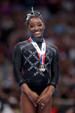 simone biles smiles at the camera, she wears a black leotard with gemstones, a white hair ribbon, and a gold medal with a red, white, and blue lanyard