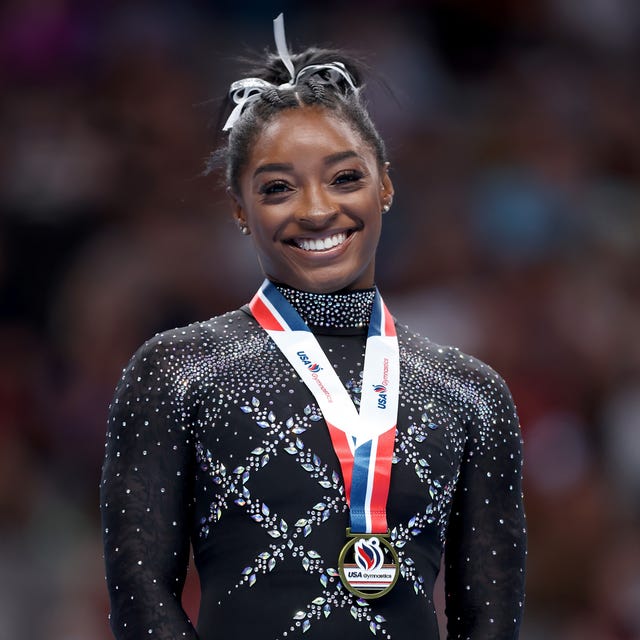 simone biles smiles at the camera, she wears a black leotard with gemstones, a white hair ribbon, and a gold medal with a red, white, and blue lanyard