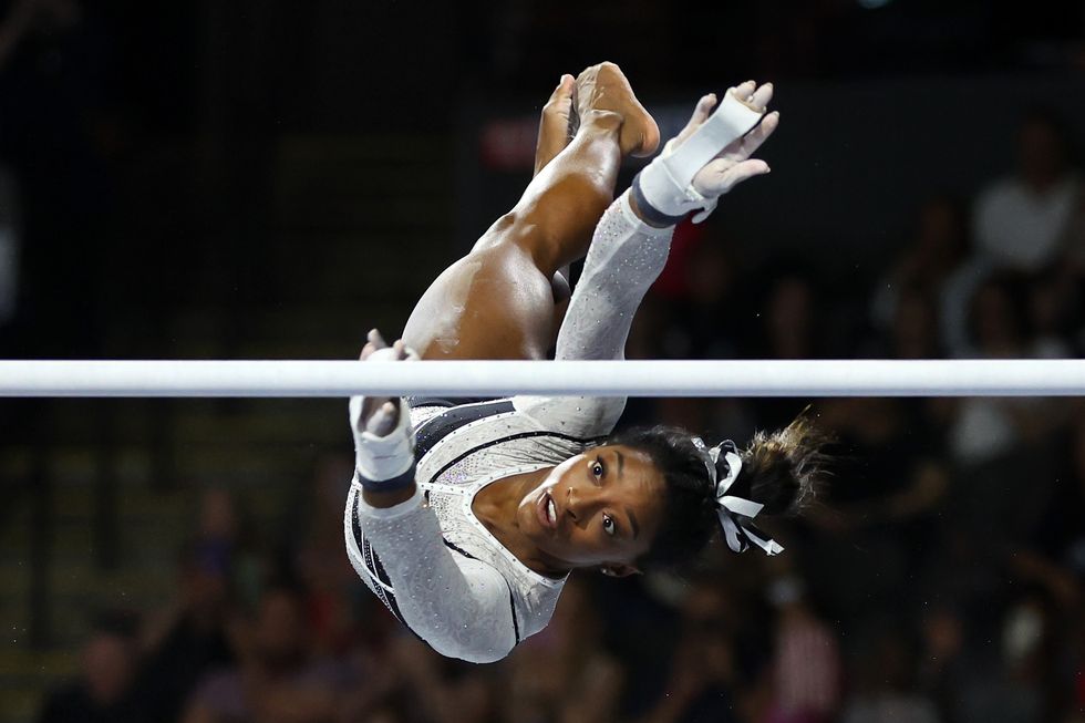simone biles flies in the air and twists to reach for an uneven bar, she wears a white and black bejeweled leotard with a matching bow in her hair