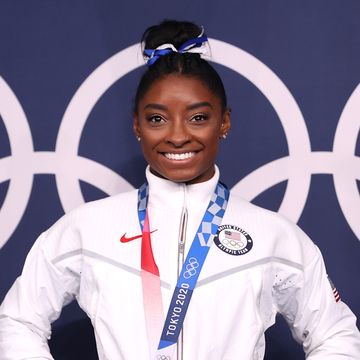 simone biles smiles while standing in front of an olympic ring logo, she is wearing a white us team jacket and a medal around her neck