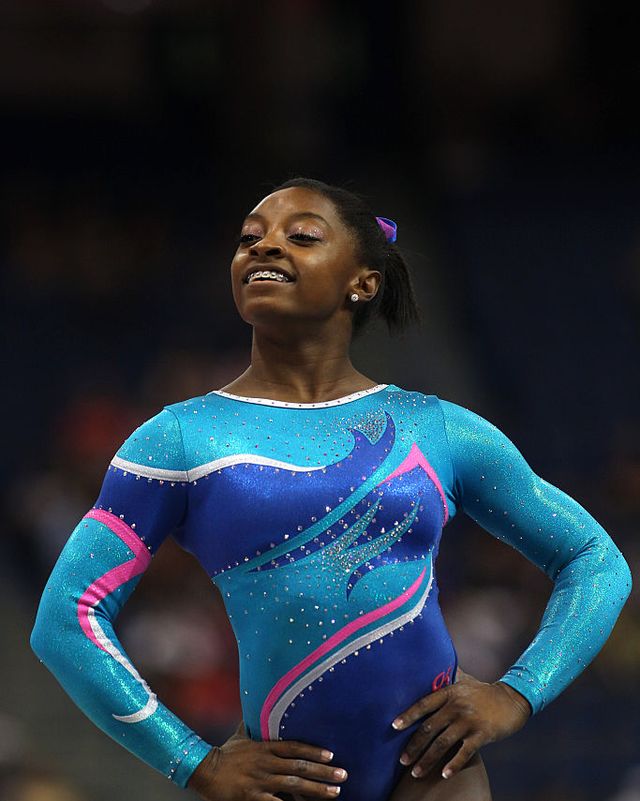 simone biles stands with her hands on her hips and raises her head slightly with a smile on her face, she wears a teal, pink, white and royal blue leotard and matching hair ribbon and has braces