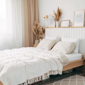 simple bulb lamp on a rope hanging above bed with white bedclothes, books and gold fern leaf on an end table in white bedroom interior
