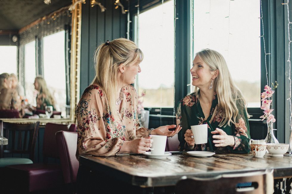 a front view shot of two beautiful mid adult women enjoying coffee together in a cafe, they are sitting at a table and laughing with eachother
