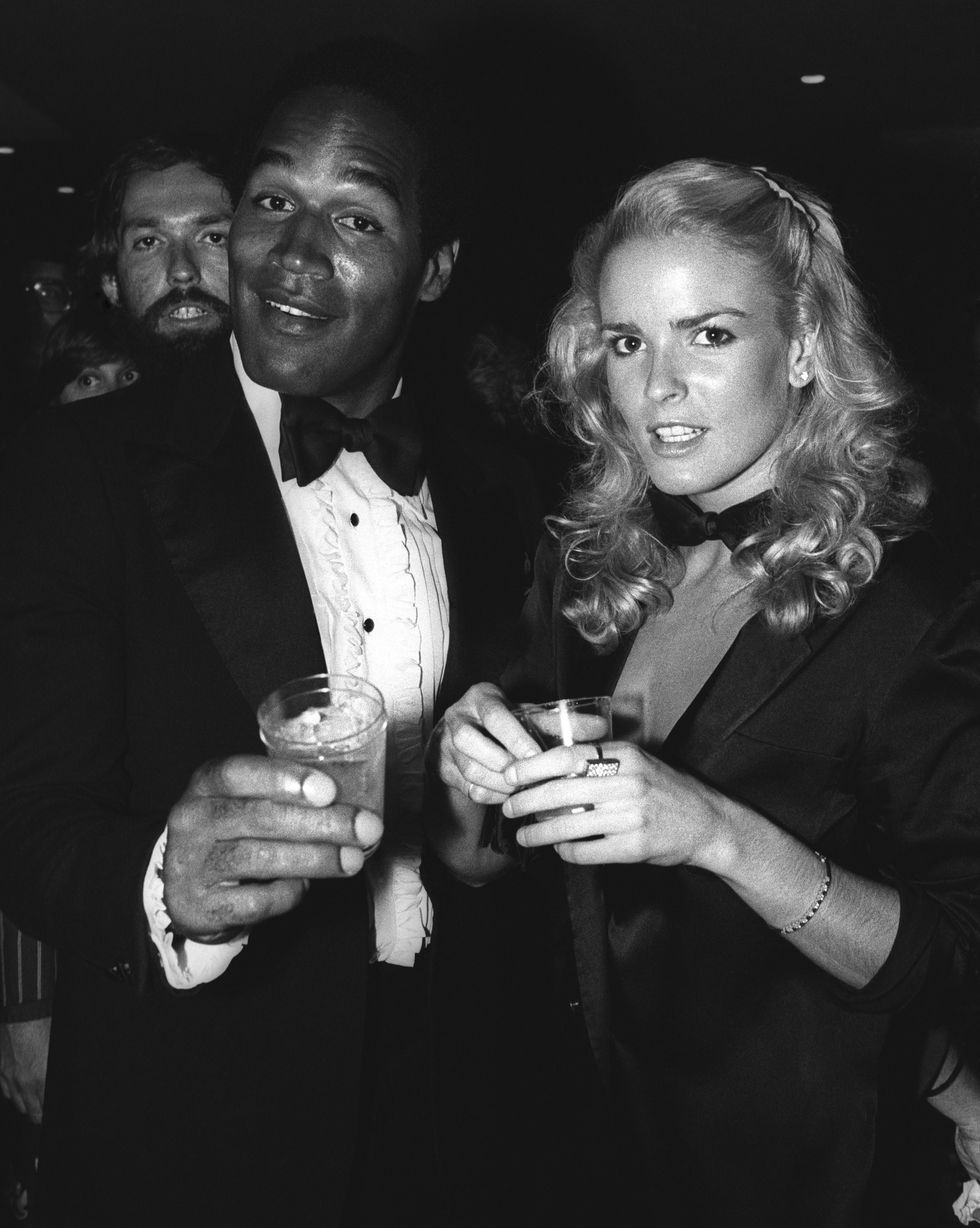 oj simpson and nicole brown look at the camera and each hold a drink, he wears a tuxedo with a bowtie and frilled shirt, she wears a suit jacket and bowtie