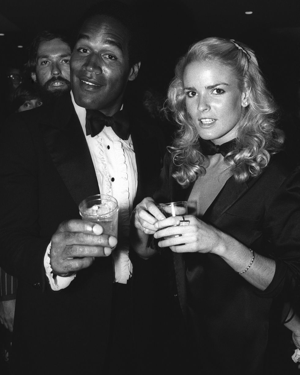 oj simpson and nicole brown look at the camera and each hold a drink, he wears a tuxedo with a bowtie and frilled shirt, she wears a suit jacket and bowtie
