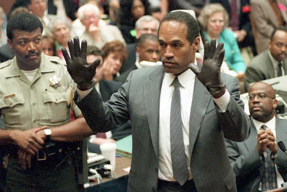 oj simpson raises both of his hands in the air in front of him and looks left, he wears black gloves and a gray suit and tie with a collared white shirt, many people look on from the background