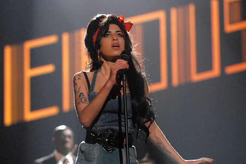 amy winehouse holding a standing microphone and singing as her name is illuminated in the background