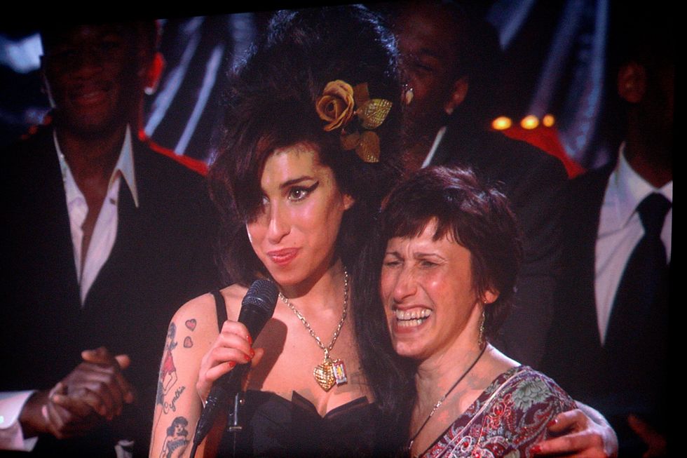 amy winehouse hugs her mother with one arm and holds a microphone on a stand in the other, both women smile as they stand on stage with a few men in suits in the background
