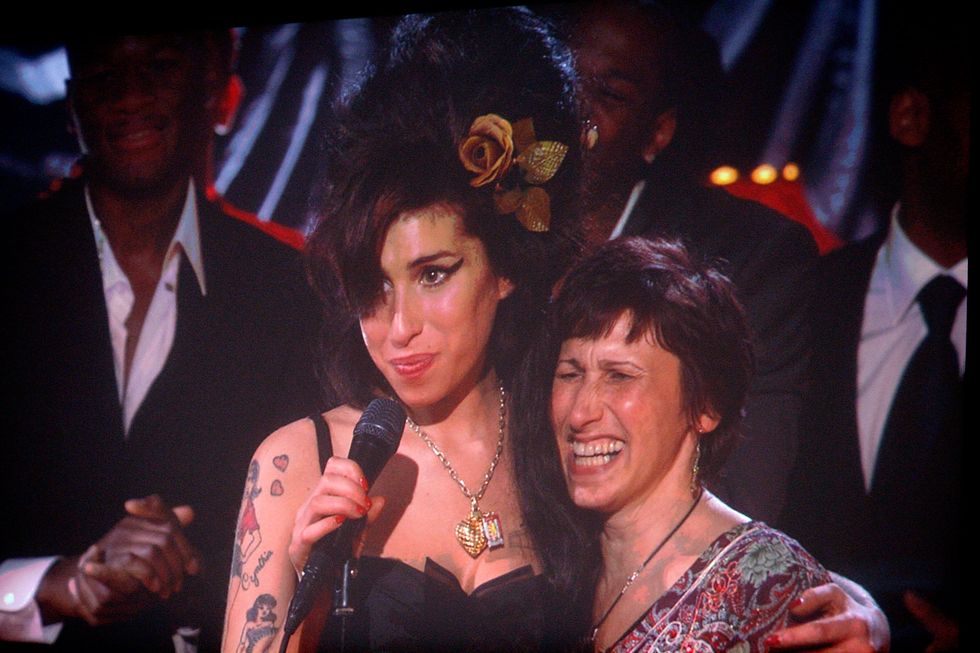 amy winehouse hugs her mother with one arm and holds a microphone on a stand in the other, both women smile as they stand on stage with a few men in suits in the background