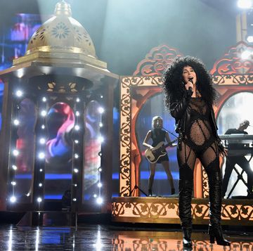 cher sings into a microphone she holds in one hand, she stands on a stage with props and musicians playing behind her