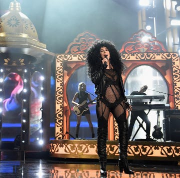 cher sings into a microphone she holds in one hand, she stands on a stage with props and musicians playing behind her