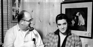 colonel tom parker looks at a smiling elvis presley while the men are seated inside a room, parker has a pipe in his mouth and a typewriter in front of him, pictures hang on the wall around them