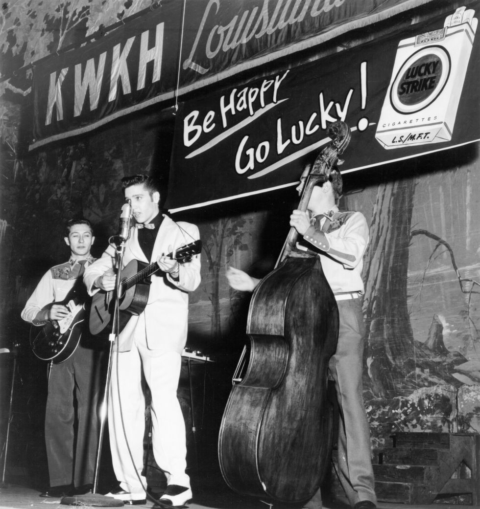 three musicians play instruments while standing on a stage, the man in the middle sings into a microphone on a stand