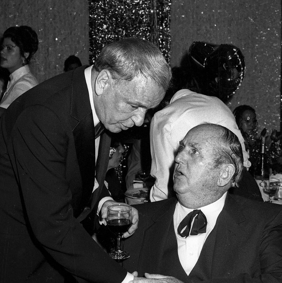 a man leans down to another man sitting at a table, they shake hands and talk, both men wear formal suits