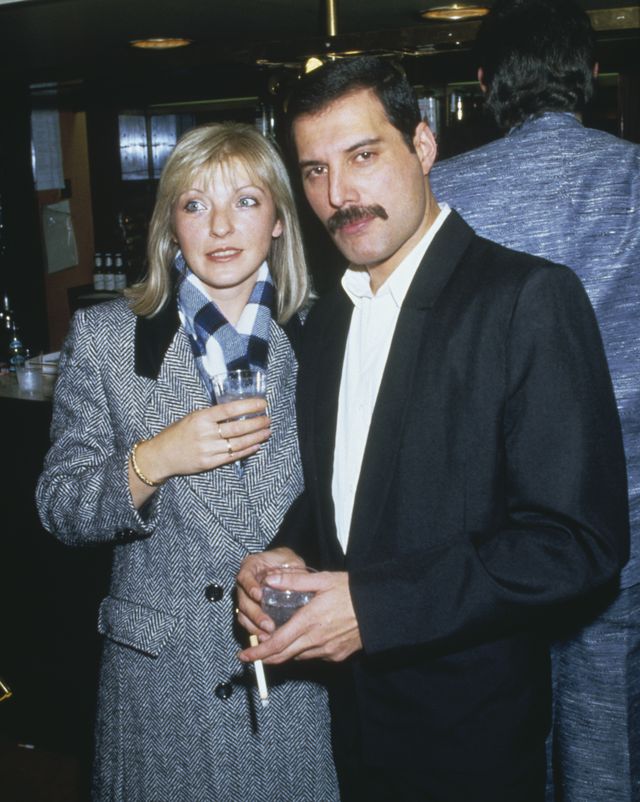 mary austin and freddie mercury stand next to each other at an event, both are holding drinks and mercury has a cigarette in one hand, she is wearing a chevron black and white jacket with a plaid scarf, he is wearing a black suit without a tie