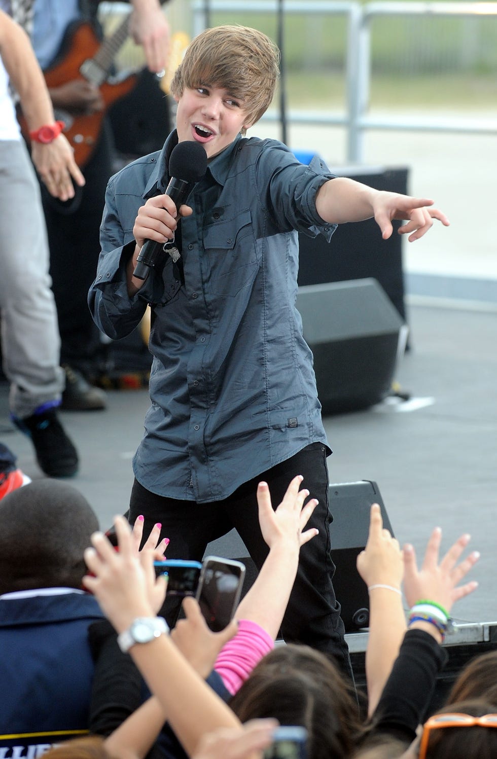 justin bieber stands on a stage and sings into a microphone he holds in one hand as he points forward with the other hand, fans stand beside the stage and reach up to him and take pictures