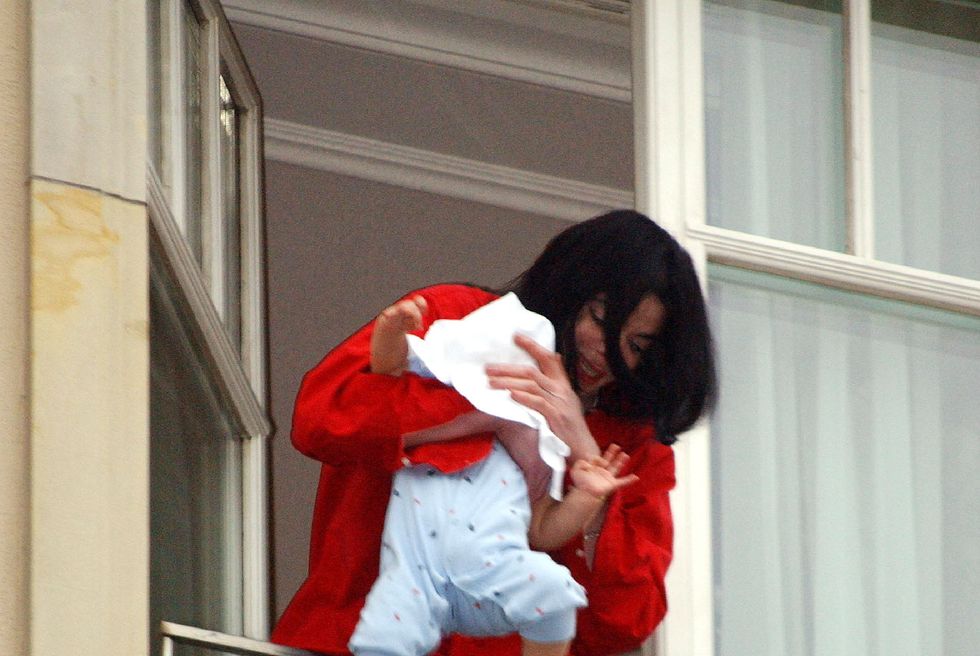 michael jackson stands on a balcony in a red long sleeve shirt and black pants, he holds a baby in a blue onesie over a railing and covers the babys face with a white piece of cloth
