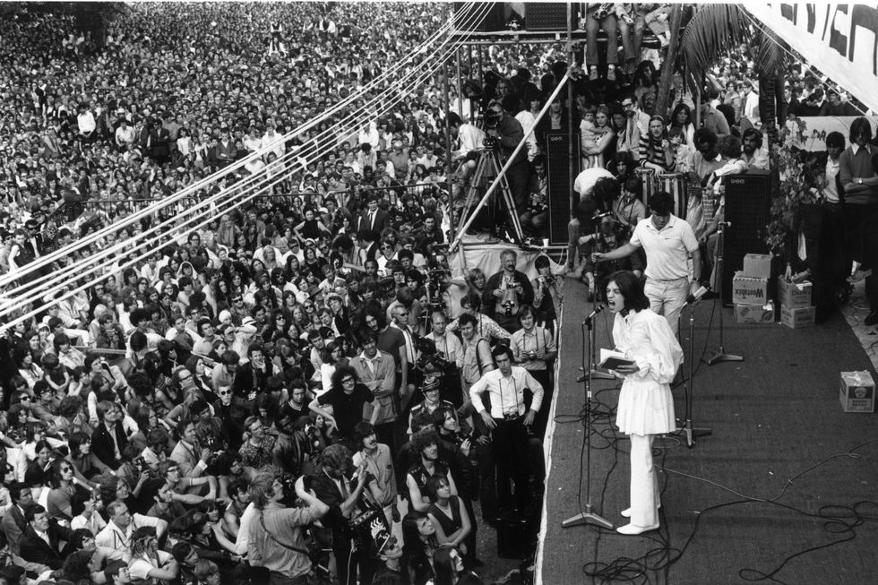 mick jagger speaks into a microphone while standing on a stage, a huge crowd watches on the left as a few other people stand on the stage