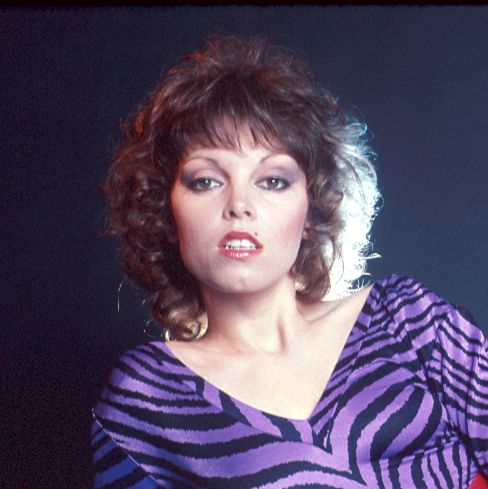 pat benatar looks directly at the camera, she wears a purple and black zebra print top, red lipstick, and purple eyeshadow, her shoulder length hair is curled