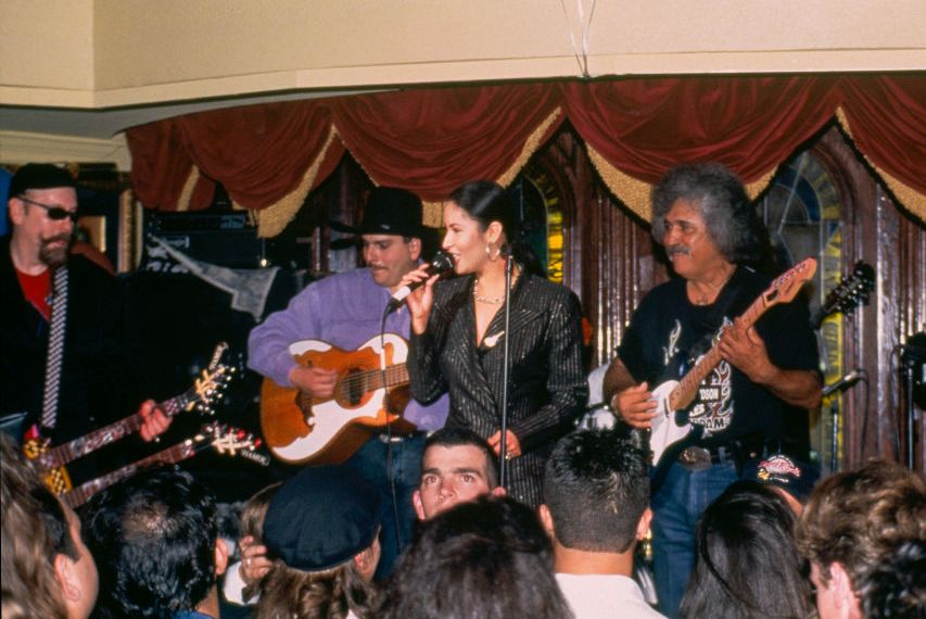 selena quintanilla sings into a microphone she holds as she stands on a stage with three musicians behind her, a crowd of people sits in front of the stage