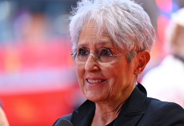 a closeup of joan baez, wearing a black shit and glasses, smiling and looking off camera, a faded background behind her