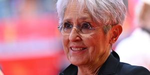 a closeup of joan baez, wearing a black shit and glasses, smiling and looking off camera, a faded background behind her