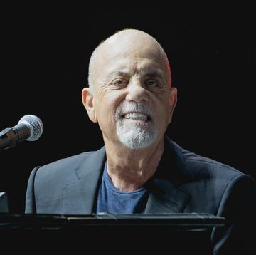 billy joel smiles at the camera as he sits at a piano, he wears a dark suit jacket and navy blue tshirt