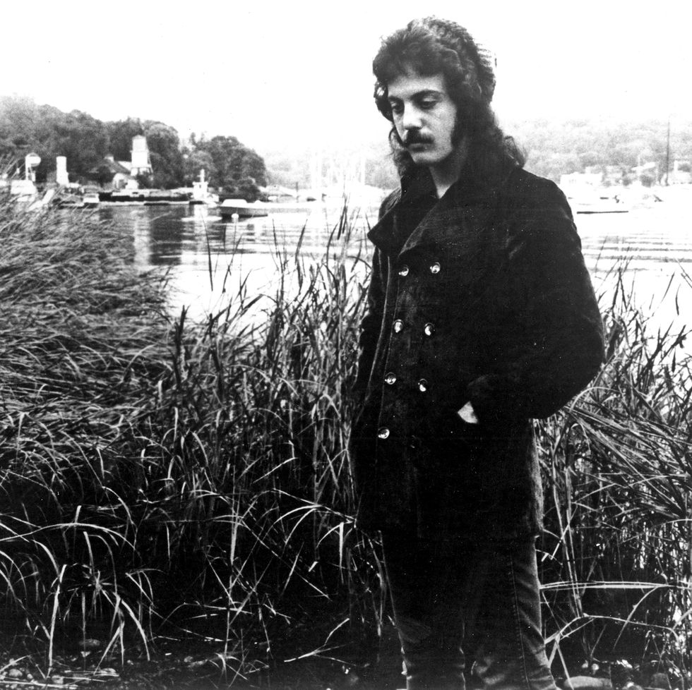 a man in a peacoat stands at a riverbank and stares at the ground facing away from the water, tall grasses boats and buildings dot the background