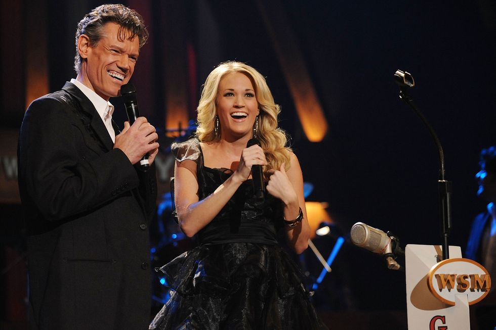 randy travis and carrie underwood smile and stand on a stage while holding microphones near their faces, he wears a black suit jacket and white collared shirt, she wears a black tulle dress and large silver hoop earrings