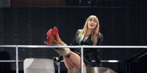 taylor swift smiles while sitting on a wooden desk with her legs extended and feet resting on a metal railing, to the left is a white office chair