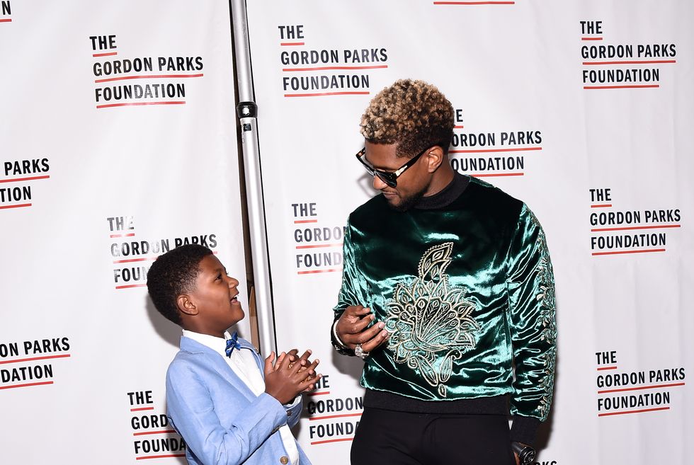 usher, wearing a blue and black designed shirt, and his son naviyd ely, wearing a blue tuxedo, speak in front of a promotional wall for the gordon parks foundation