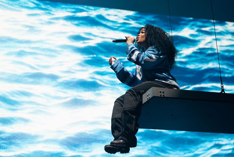 sza sits on the end of a hanging platform with her legs hanging off the end, she sings into a microphone she holds in one hand, behind her is a large screen projecting waves, she wears a long sleeve blue jersey style shirt, black cargo pants, and black boots