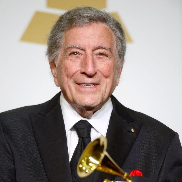 tony bennett smiles for a photo, he wears a black suit and tie with a red pocket square, part of a grammy award is visible in the foreground