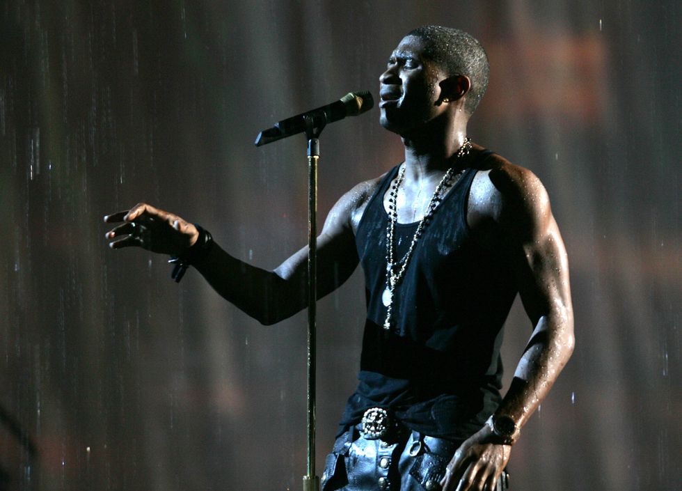 usher sings into a microphone on a stand while reaching one hand in front of him, he wears a black tank top, jeans, two necklaces, and a watch, water rains down on him