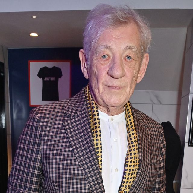 ian mckellen looks at the camera with a small smile, he wears a white button up shirt, brown and tan plaid suit jacket and yellow scarf with a blue circle pattern