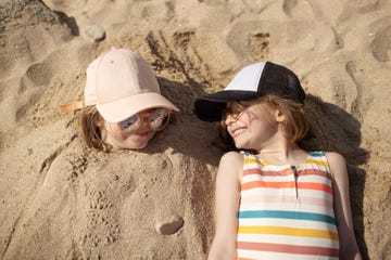 six year old girls laughing together on the beach, one girl has her body buried in the sand and the other girl is laying next to her friend