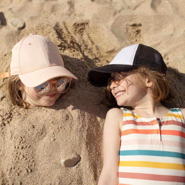 six year old girls laughing together on the beach, one girl has her body buried in the sand and the other girl is laying next to her friend