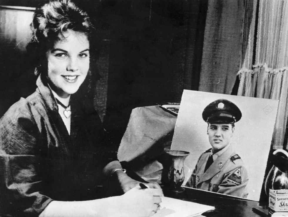 priscilla beaulieu sits at a table, holding a pen over paper as she smiles at the camera, a square photo of elvis is propped up on the table, priscilla wears a dark shirt and a heart pendant necklace