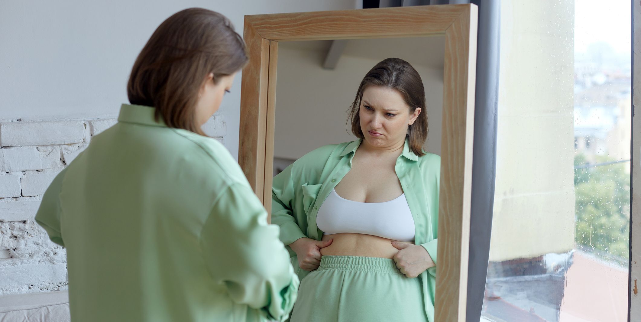 size plus woman looking by reflection in mirror feels frustrated and dissatisfied by overweight