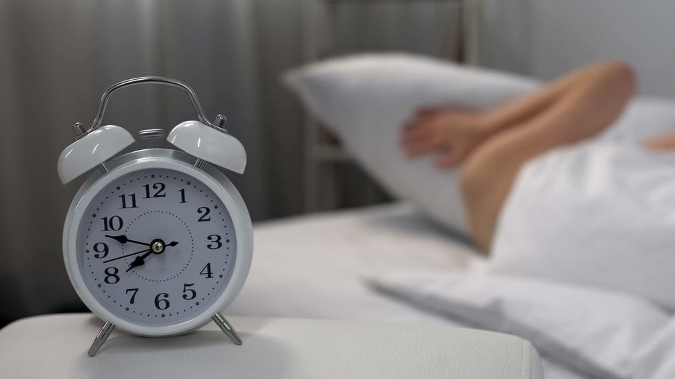 sleepy female annoyed by ringing alarm clock closing ears with pillow, jet lag