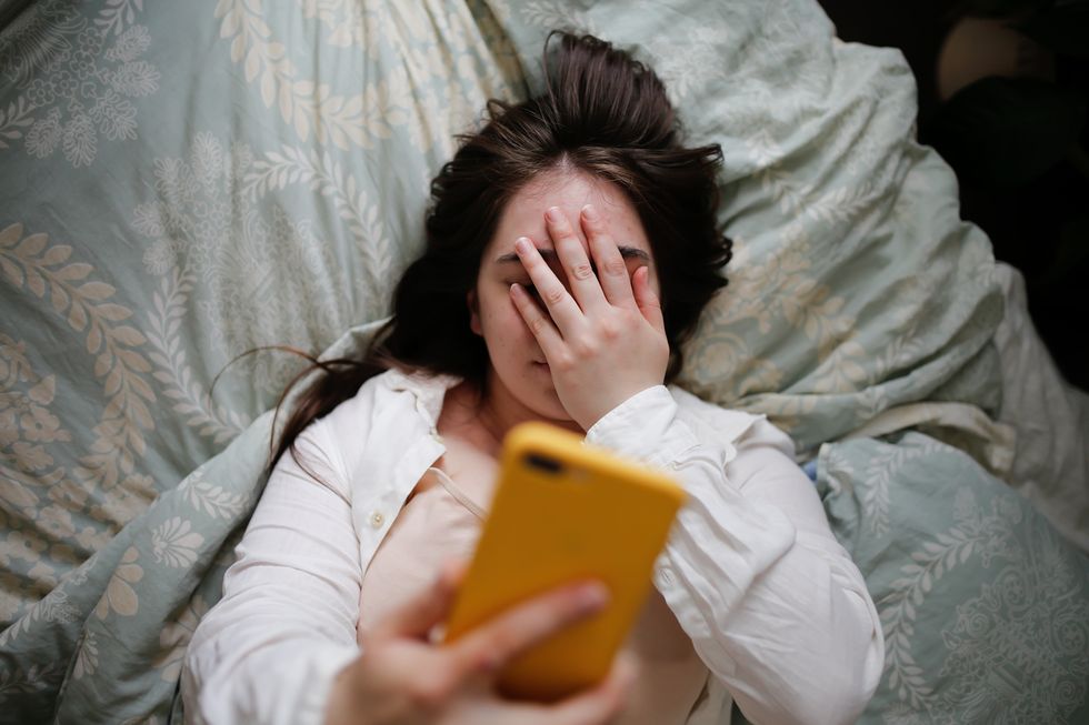 sleepy overweight young woman with flowing hair using phone on bed, close face palm top view