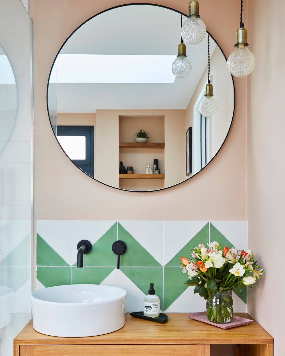 a round mirror above green and white tiles in a small bathroom painted pale pink