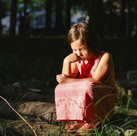a small forlorn child sits in sunlight in a shaded patch of woods