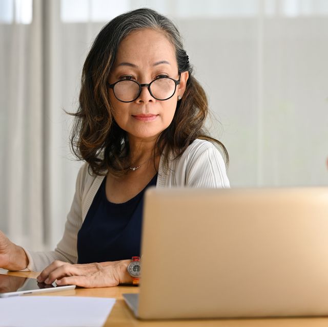 smiling 60s middle aged business lady remote working from home, using laptop watching webinar and taking notes on digital tablet.