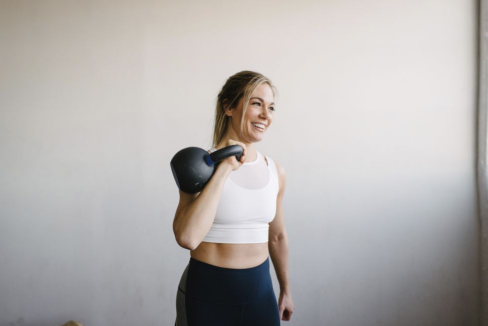 smiling female athlete carrying kettlebell while standing by wall in gym
