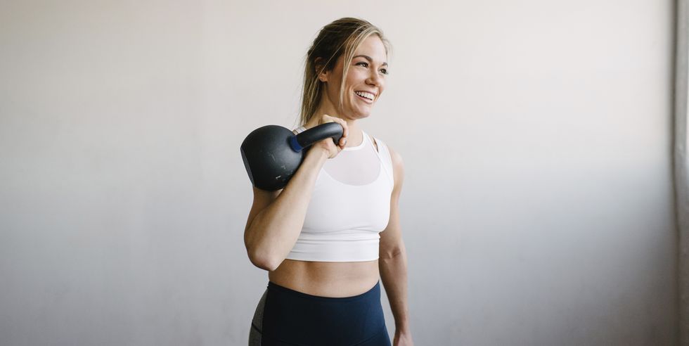 smiling female athlete carrying kettlebell while standing by wall in gym