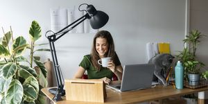 smiling female entrepreneur holding mug using digital tablet on desk in home office